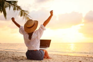 Work from anywhere. Rear view of young woman, female freelancer in straw hat working on laptop near fort myers, keeping arms raised and cellebrating success while sitting on the tropical sandy beach at sunset in front of her remote work vacation rentals with resort harbour properties